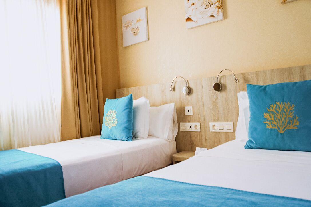 Habitación de hotel con dos camas, cada una con colcha azul y almohadas a juego. Hay enchufes en la pared y un cabecero con altavoces integrados.