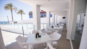 A modern outdoor dining area with white tables and chairs, overlooking a beach with palm trees and a clear sky. - Hotel Sol de Jávea (Javea)