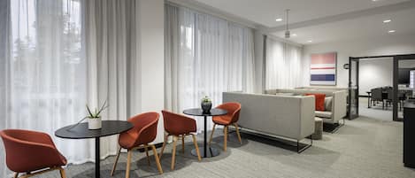 Una habitación de hotel moderna con una mesa redonda, cuatro sillas, un sofá y un ventanal con cortinas translúcidas.