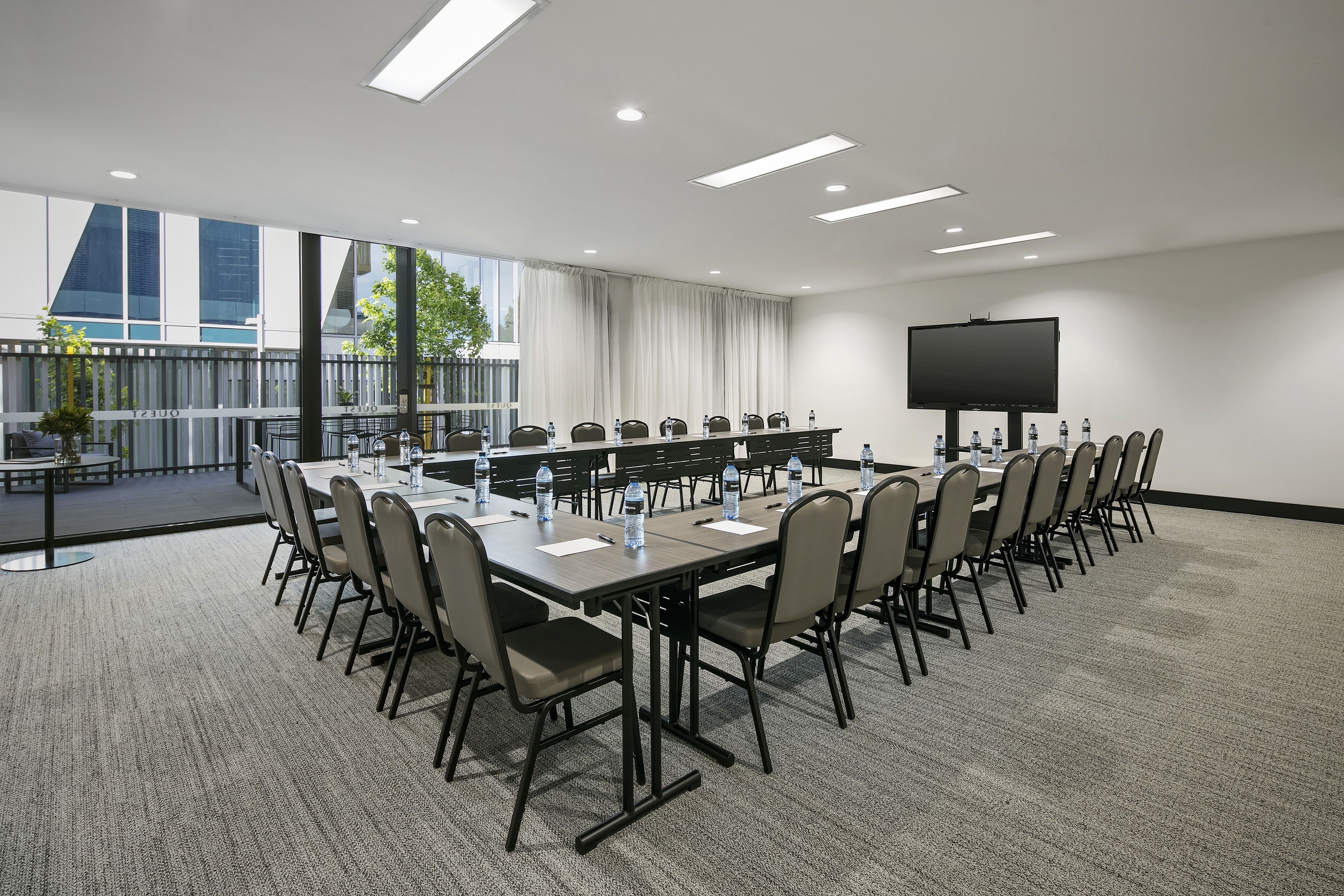 A conference room with a long table, chairs, a flat-screen TV, and a view of buildings through large windows.