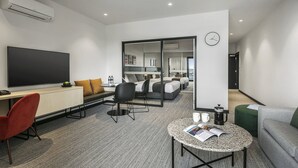 A modern hotel room with a flat-screen TV, a desk with a chair, a sofa, a coffee table, and a bed with a view of the kitchen and bathroom. - Quest Notting Hill (Notting Hill)