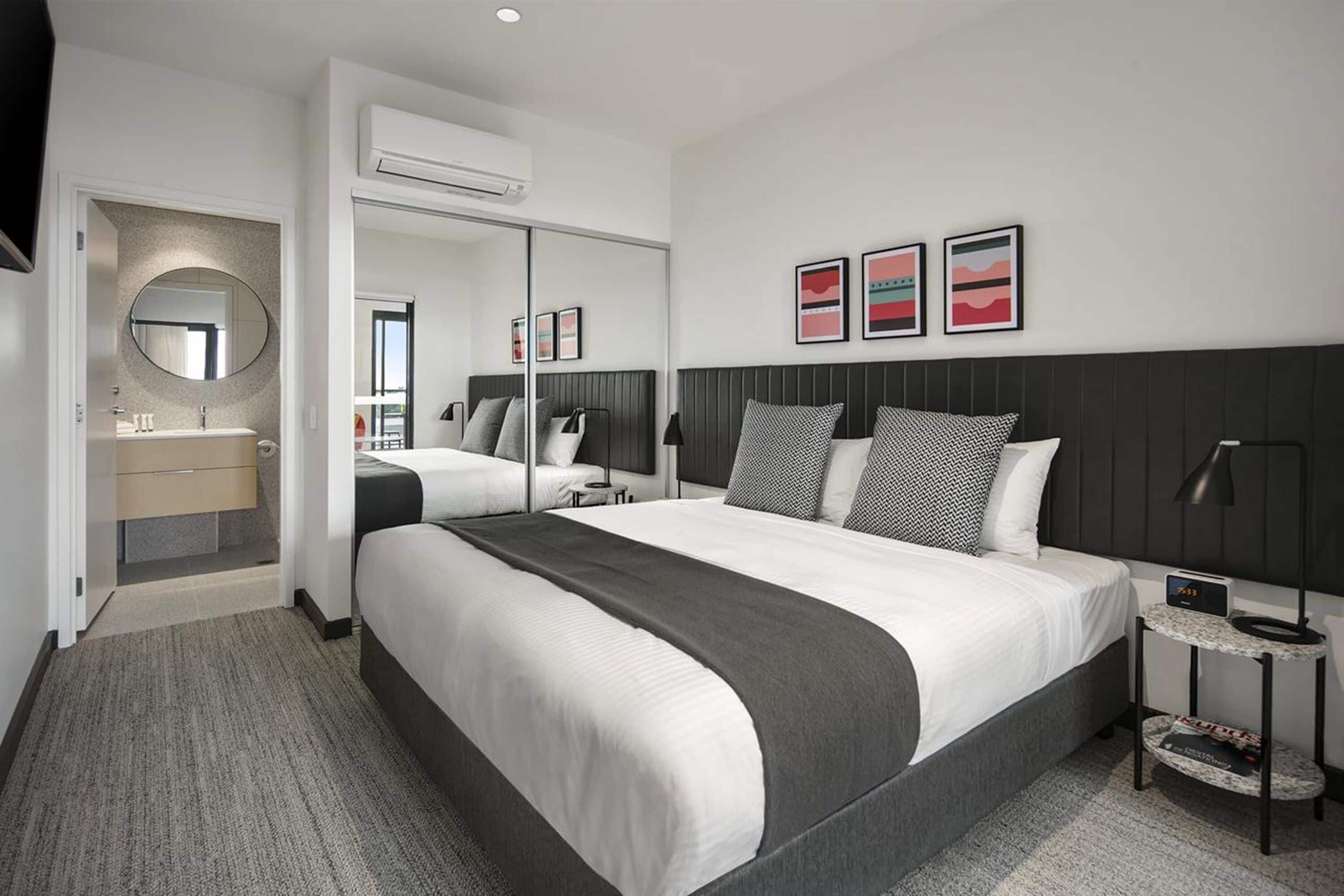 A modern hotel room with a large bed, a bedside table with a lamp, a mirror, and artwork on the wall.