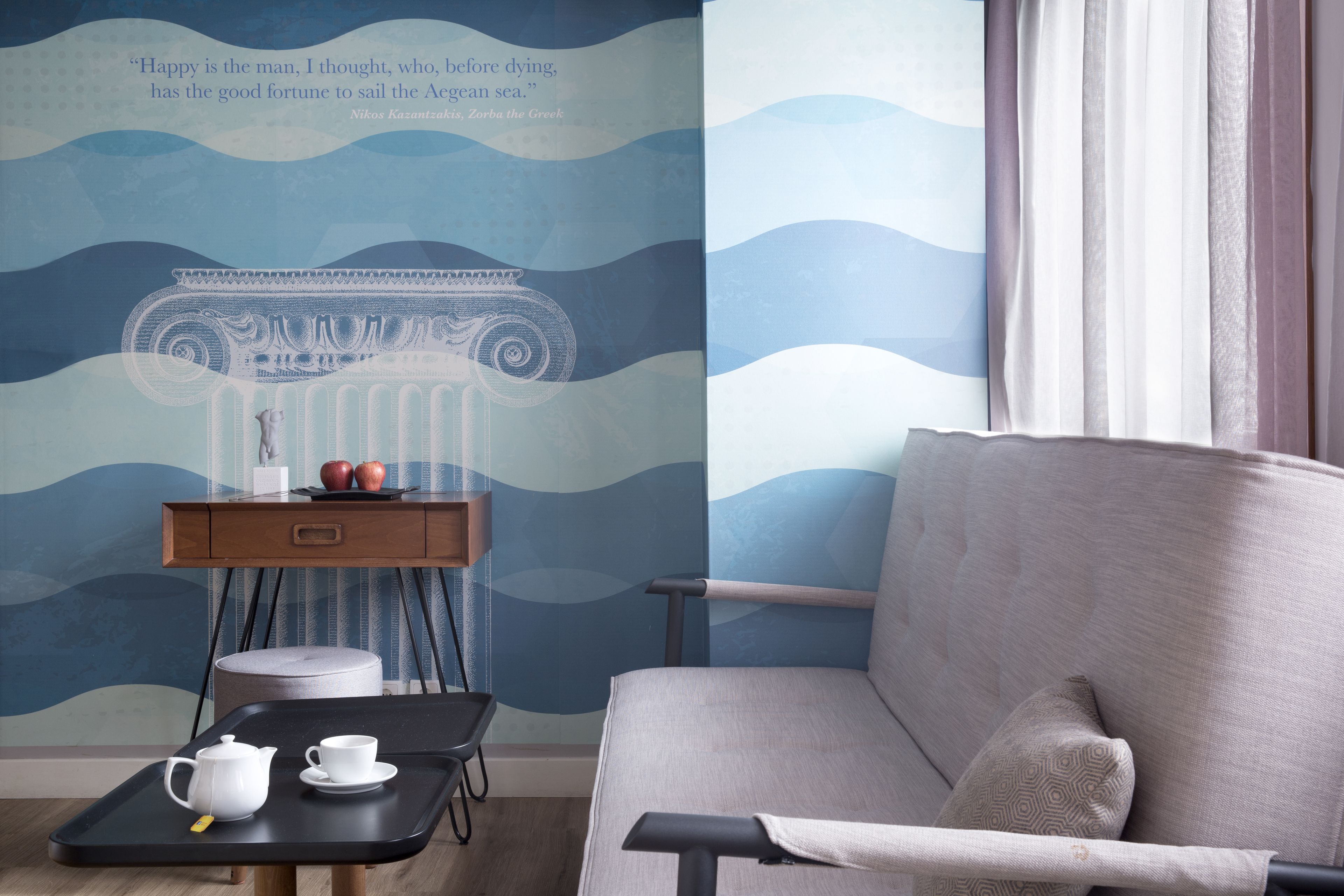 A room with a grey sofa, a small wooden side table with a tray holding a teapot and cup, and a wall with a wave pattern and a quote.