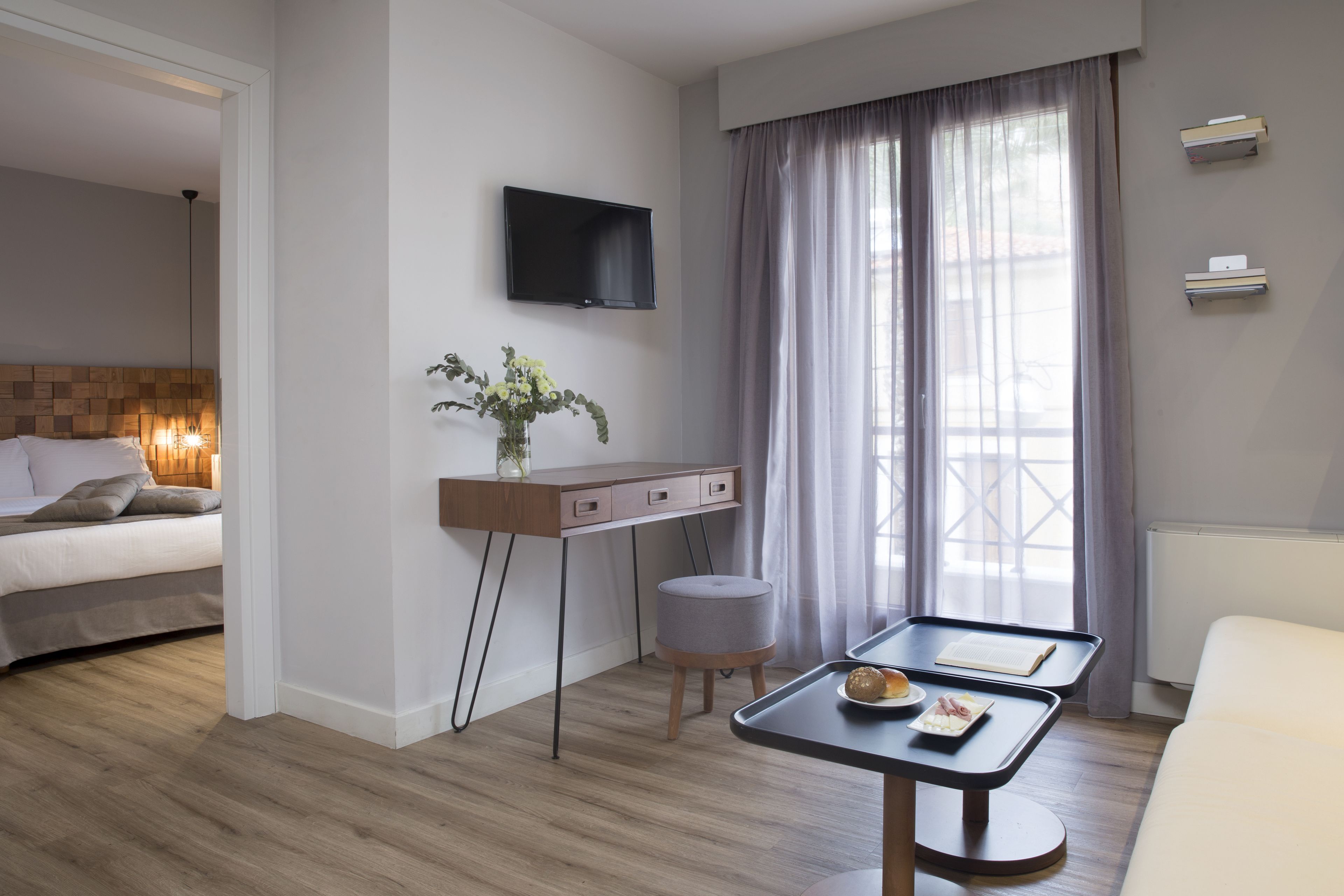 Quarto de hotel moderno com cama, escrivaninha com televisão de tela plana, pequena mesa com bandeja e uma grande janela com cortinas.