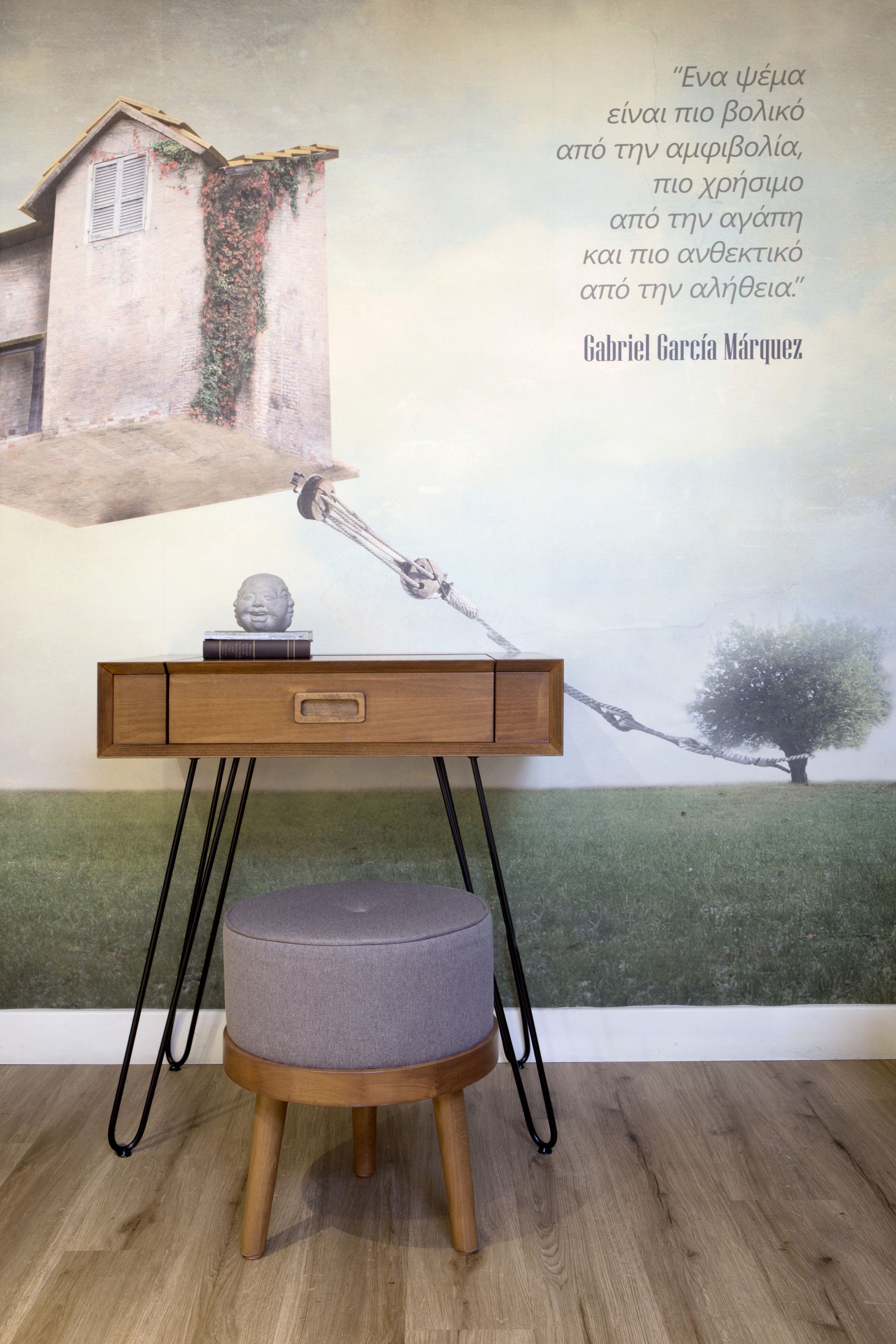 A room with a wooden desk, a grey ottoman, and a wall with a quote and a painting.