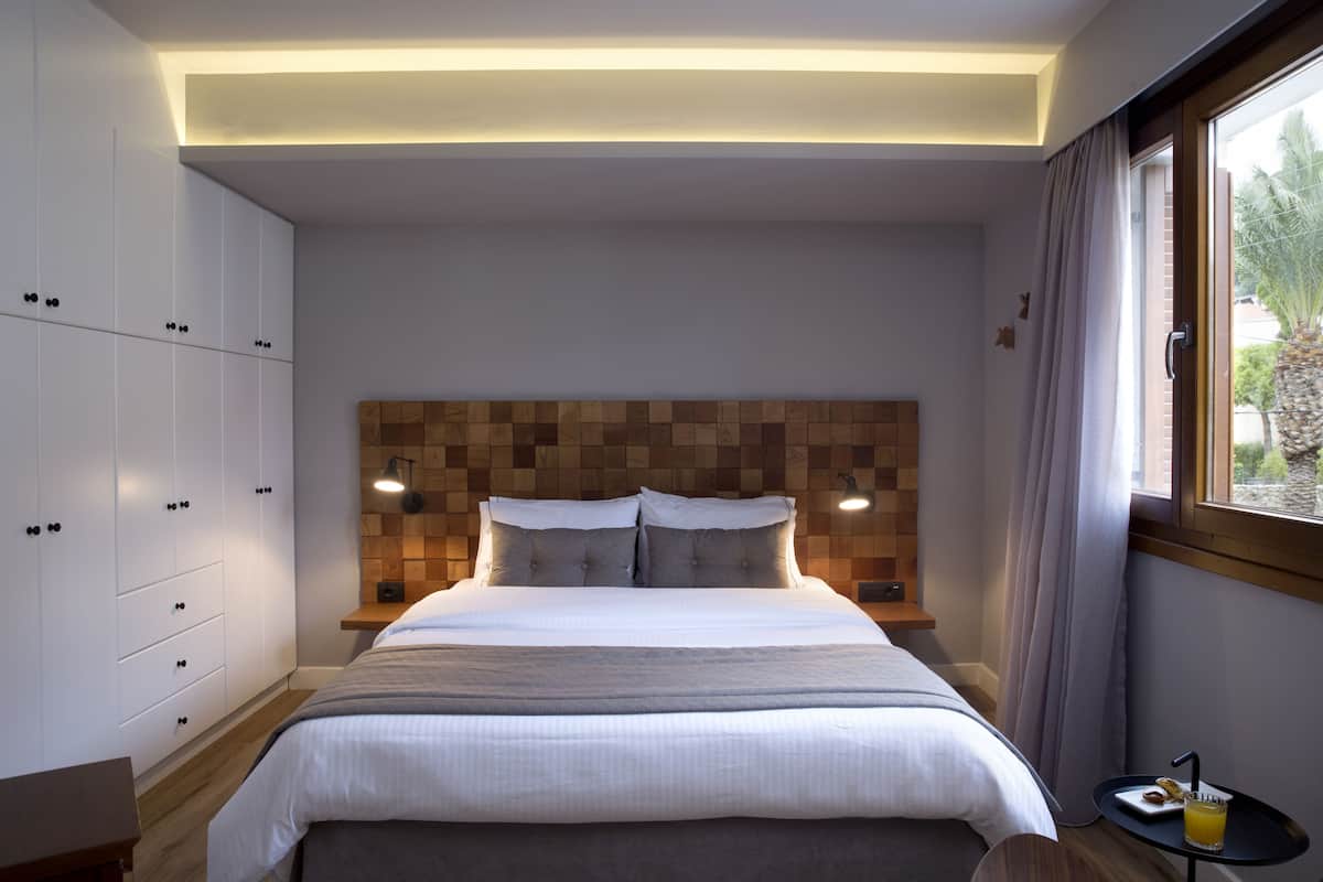 A modern bedroom with a large bed, wooden headboard, built-in wardrobe, bedside lamps, and a small table with a drink.