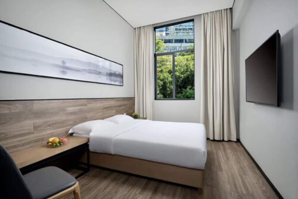 A modern hotel room with a bed, a desk with a chair, a flat-screen TV, and a large window with a view of greenery.