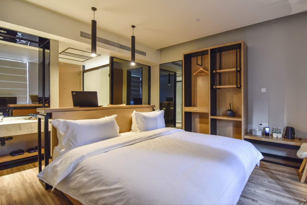 A modern hotel room with a large bed, a desk with a laptop, a chair, a wardrobe, and a mirror.