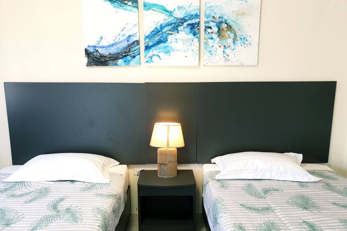 A hotel room with two beds, each with a white pillow and a leaf-patterned duvet. There is a nightstand with a lamp between the beds, and a wall-mounted artwork above the artwork.
