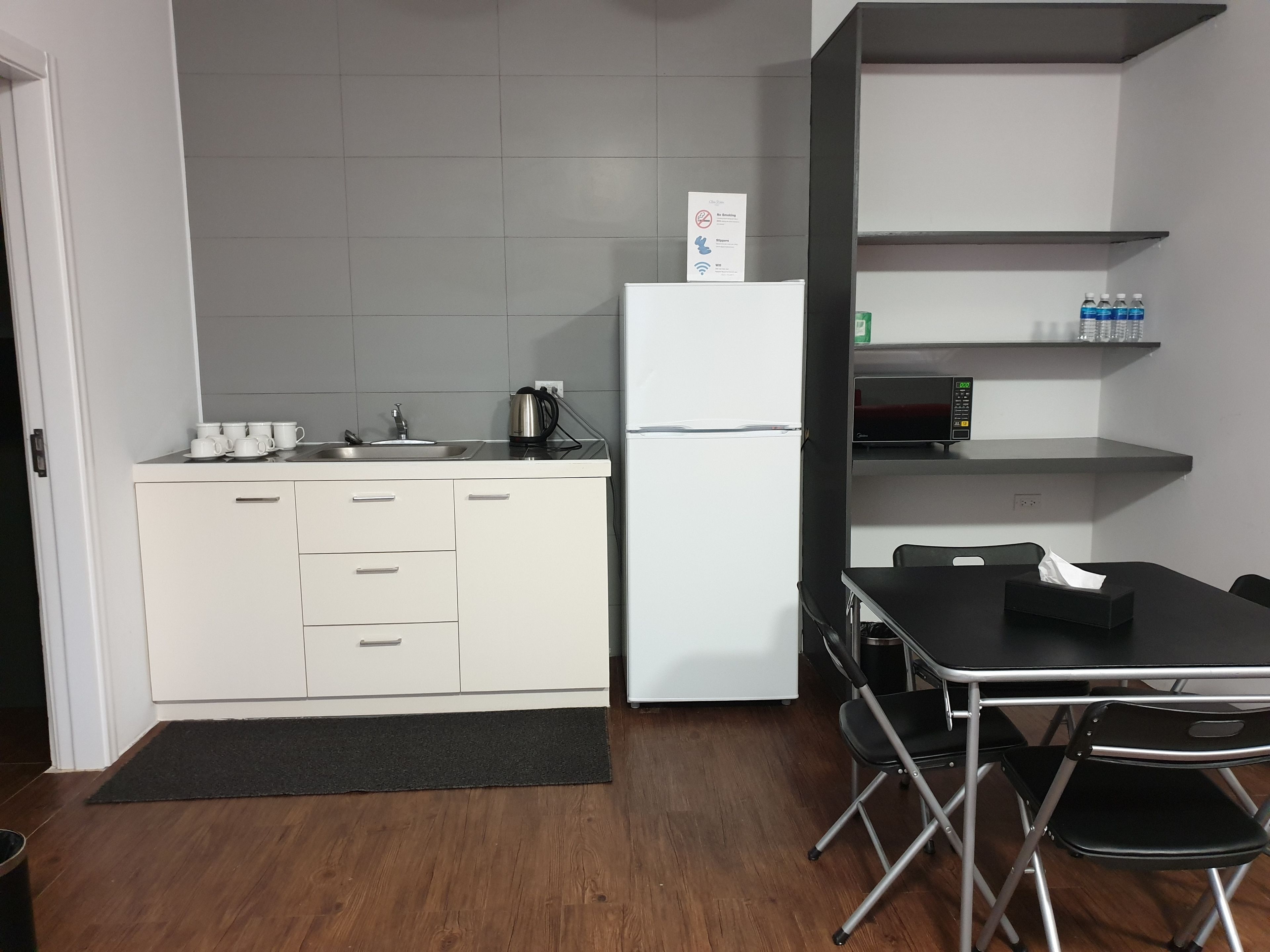 A compact kitchen with white cabinets, a stainless steel sink, and a refrigerator. There is a black table with chairs and a black mat on the floor.