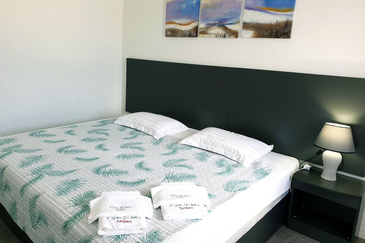 A neatly made bed with a leaf-patterned bedspread, two pillows, and two folded towels on the bed. There is a nightstand with a lamp beside the bed.