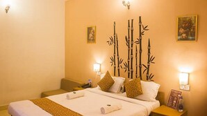 A hotel room with a bed, two bedside tables, a nightstand, a wall-mounted lamp, and a decorative bamboo wall panel. - Jesus Holiday Home Resort (Candolim)