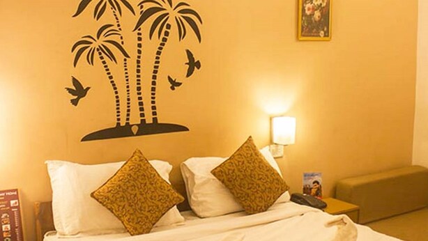 A hotel room with a bed, two pillows, a nightstand, a lamp, and a wall decoration of palm trees and birds. - Jesus Holiday Home Resort (Candolim)
