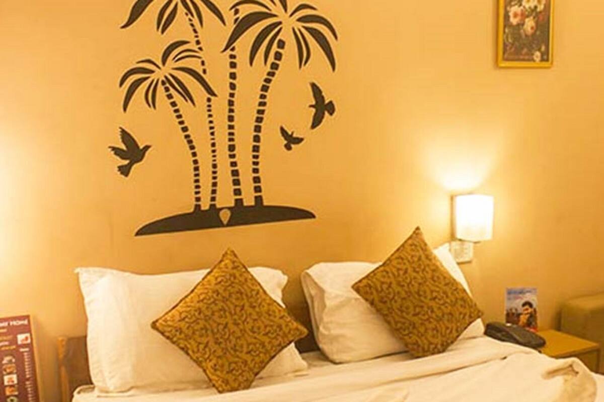 A hotel room with a bed, two pillows, a nightstand, a lamp, and a wall decoration of palm trees and birds.