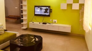 Ein Wohnzimmer mit grüner Wand, an der ein Flachbildfernseher befestigt ist. Darunter befindet sich eine weiße Unterhaltungsecke und im Vordergrund ein Glastisch.