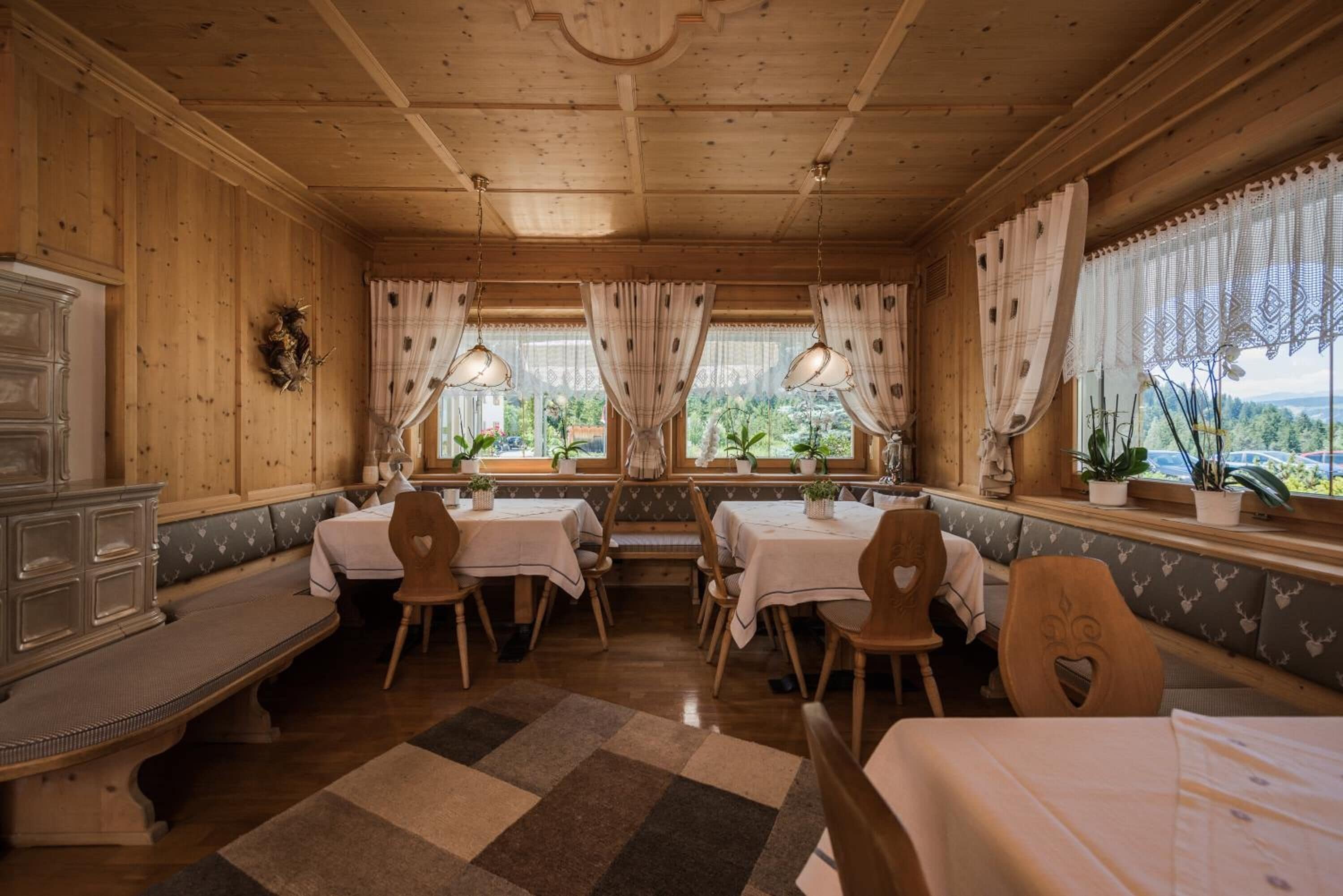 A cozy wooden room with a bench, tables, and chairs, large windows with curtains, and a view of greenery outside.