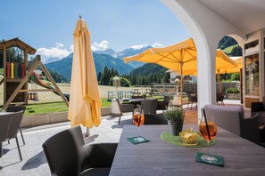 Un espacio al aire libre con mesas, sillas y sombrillas, con vistas a un parque infantil y montañas.