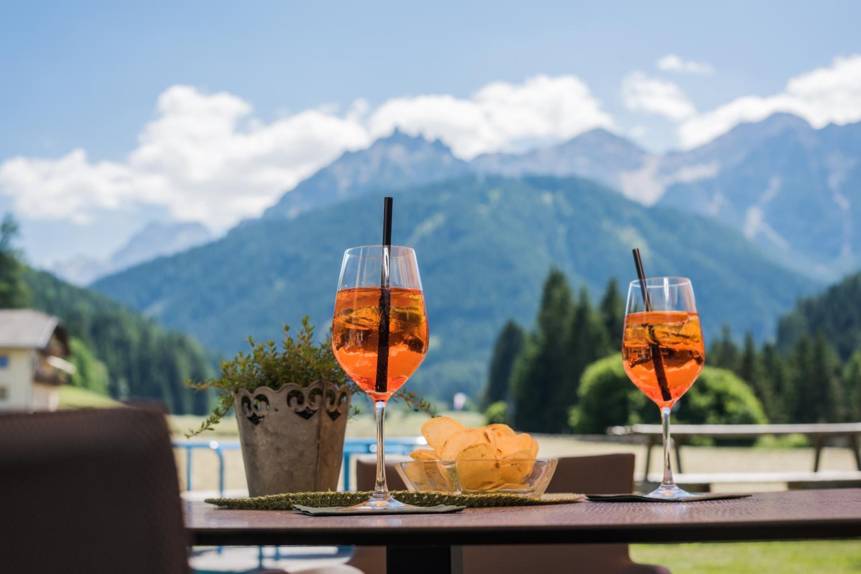 Two glasses of amber-colored drink with straws on a table with a mountain backdrop.
