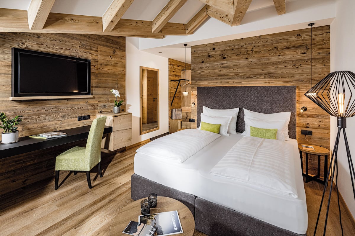 A modern hotel room with a large bed, a desk with a chair, a TV mounted on the wall, and a lamp on a side table.