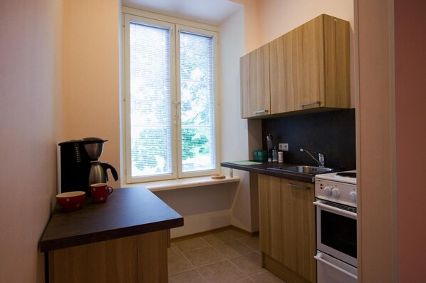 Cocina compacta con gabinetes de madera, horno empotrado, cafetera y ventana con persianas.