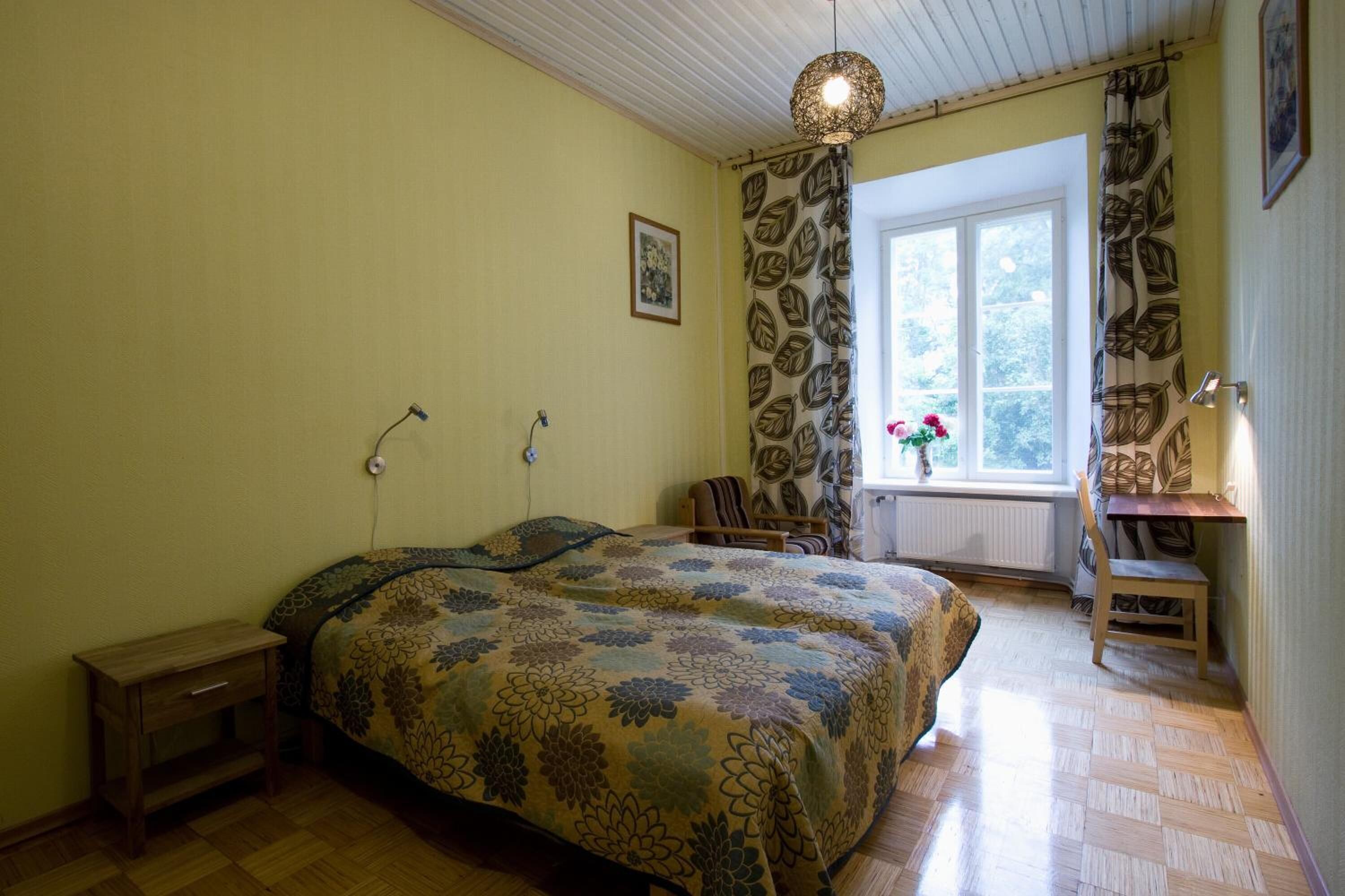 Un dormitorio con una cama, una silla, una mesita, una ventana con cortinas y una lámpara colgante.