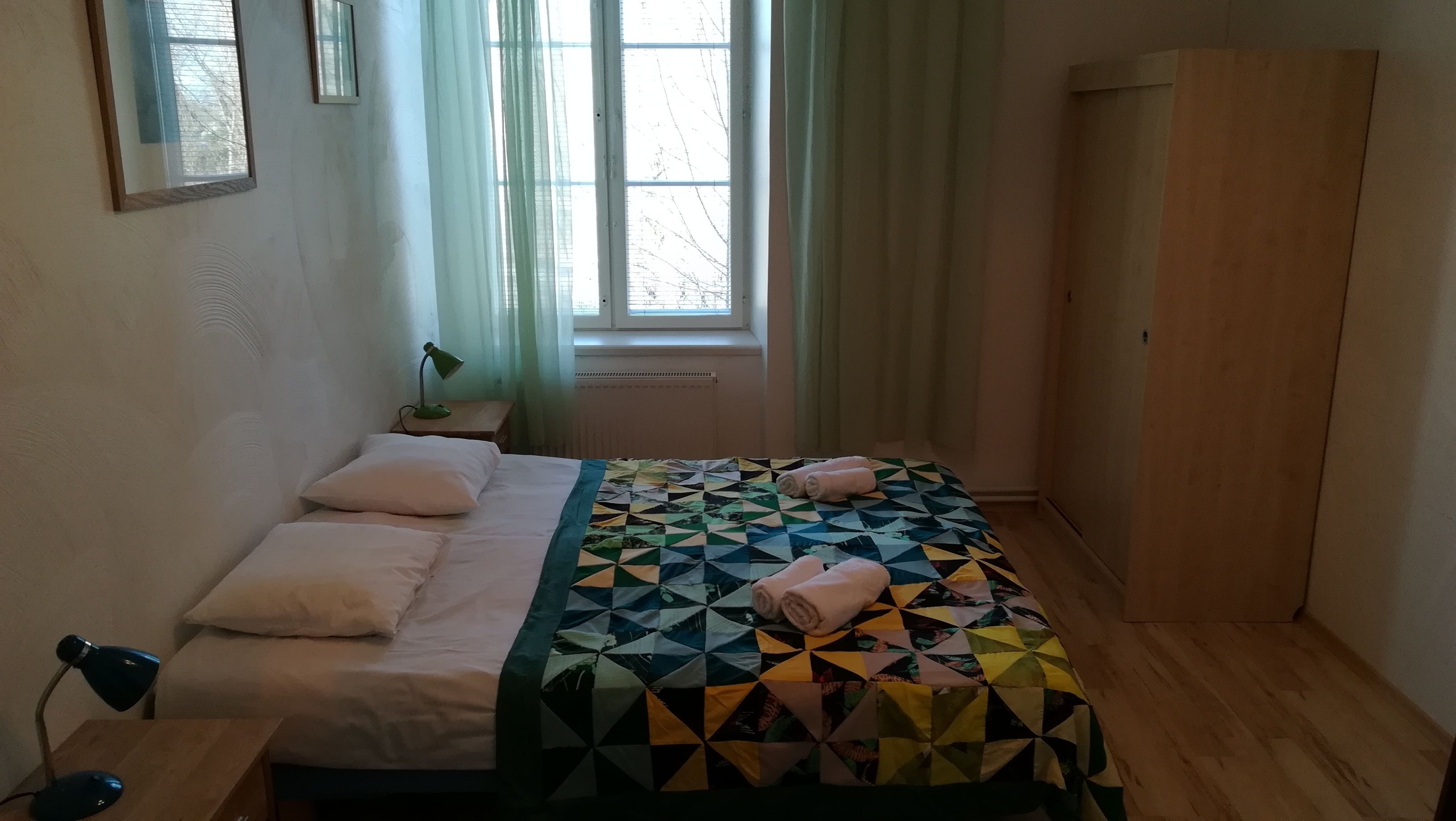 Ett sovrum med en säng, två sänglampor, ett fönster med gardiner, ett element och en garderob.