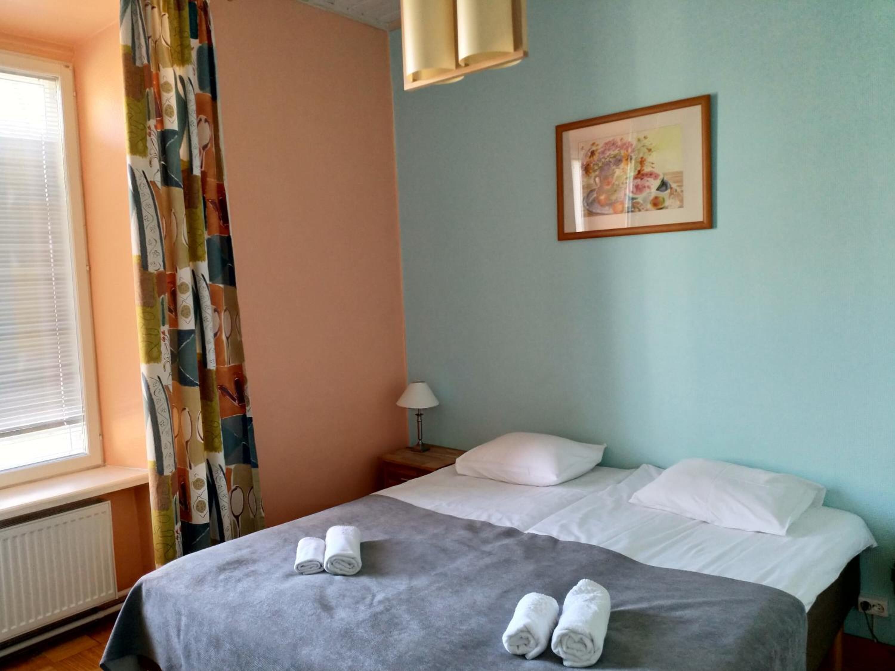 A hotel room with a bed, two rolled towels on the bed, a window with blinds, a curtain, a lamp, and a framed picture on the wall.