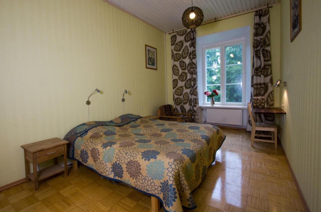 A bedroom with a bed, a small wooden nightstand, a chair, a window with curtains, and a hanging light fixture.