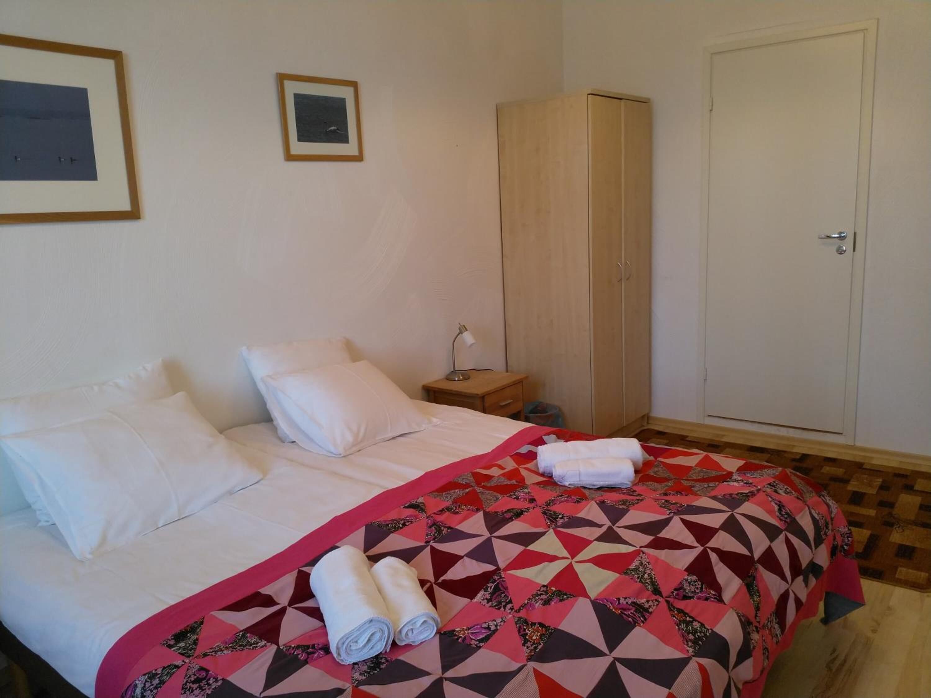 Quarto com cama, almofadas, colcha, guarda-roupa de madeira, mesa de cabeceira e uma porta.
