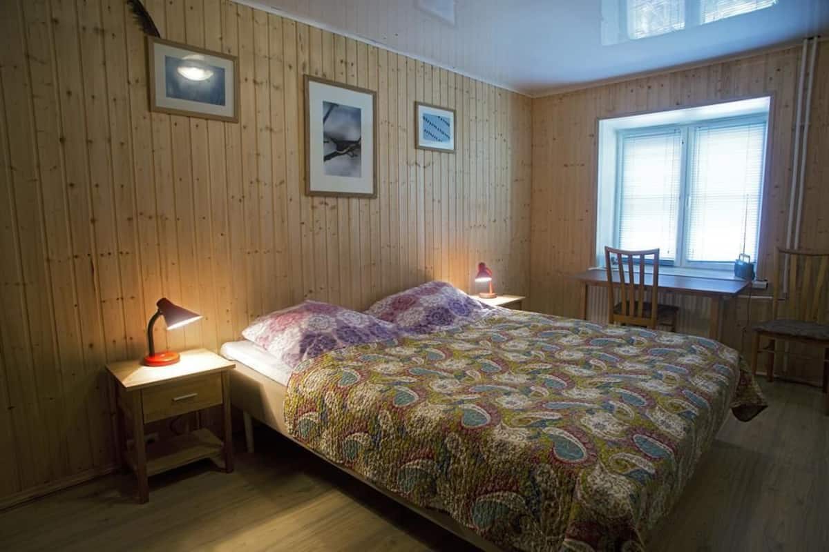 Una habitación con paneles de madera, una cama con colcha colorida, dos mesitas de noche con lámparas, una ventana con persianas y una silla junto a una pequeña mesa.