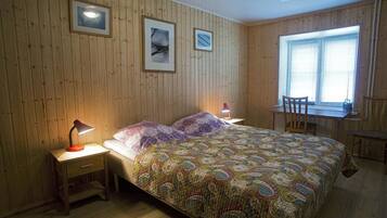 Una habitación con paneles de madera, una cama con colcha colorida, dos mesitas de noche con lámparas, una ventana con persianas y una silla junto a una pequeña mesa.