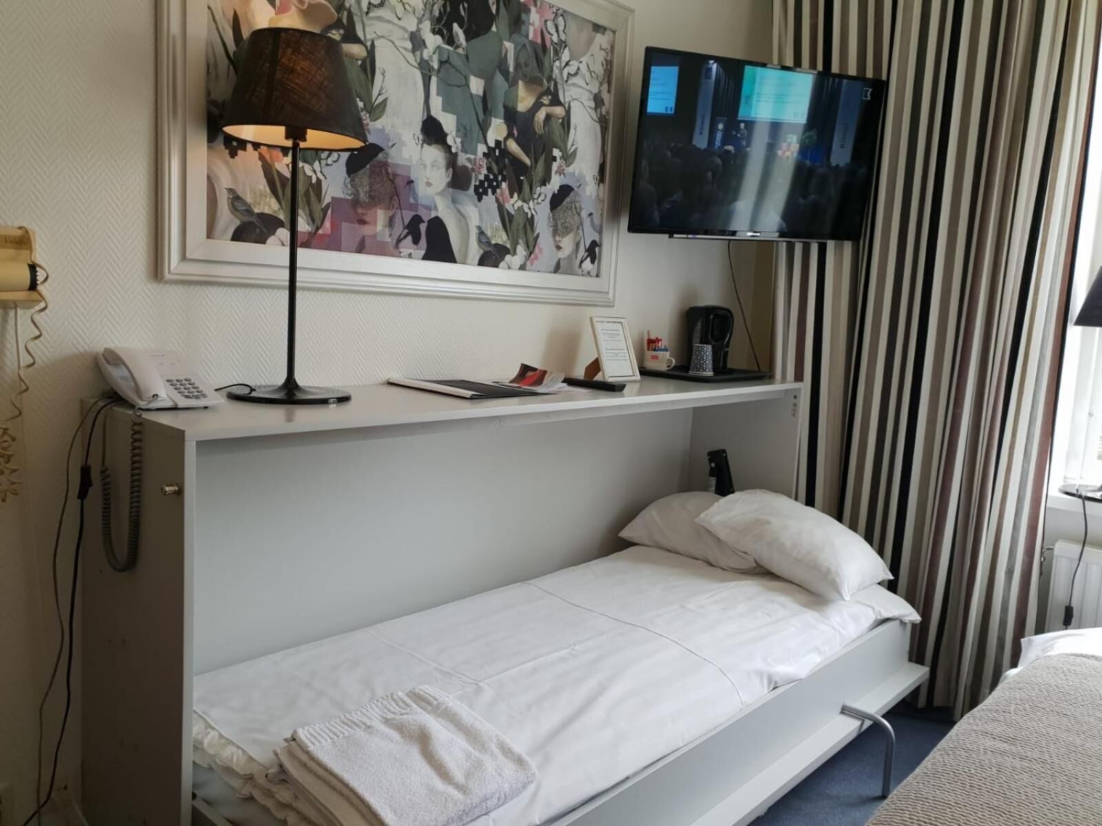 Habitación de hotel con cama, escritorio con lámpara, teléfono, televisión y un cuadro en la pared.