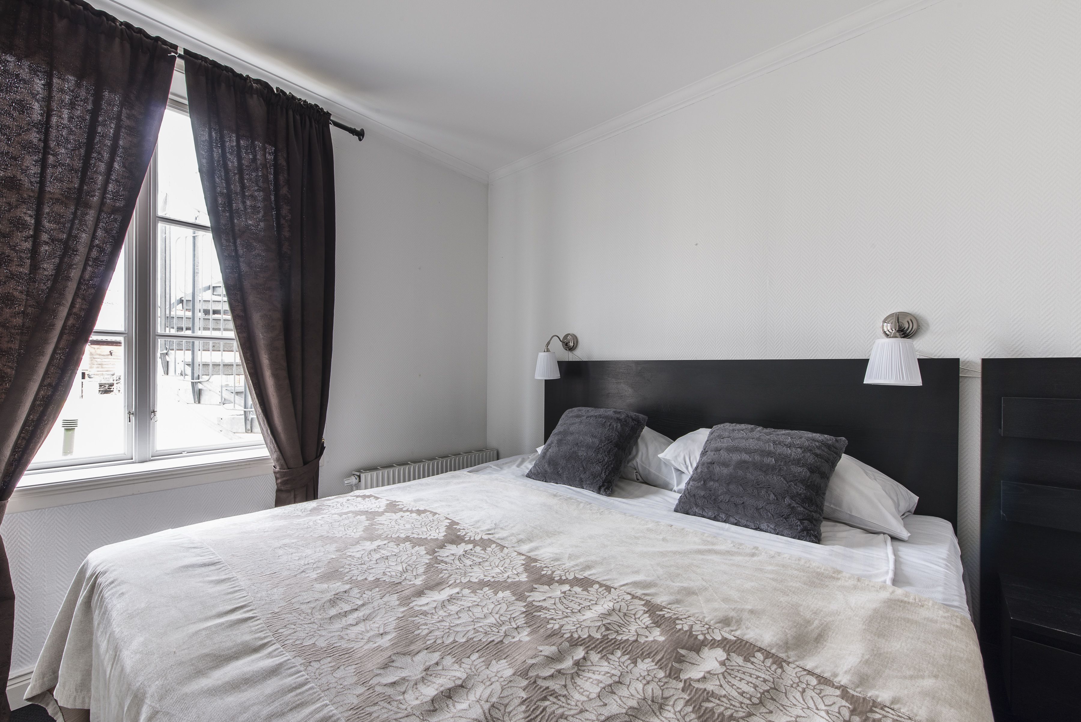 A bedroom with a large bed, two bedside lamps, and a view of the city through a window with curtains.