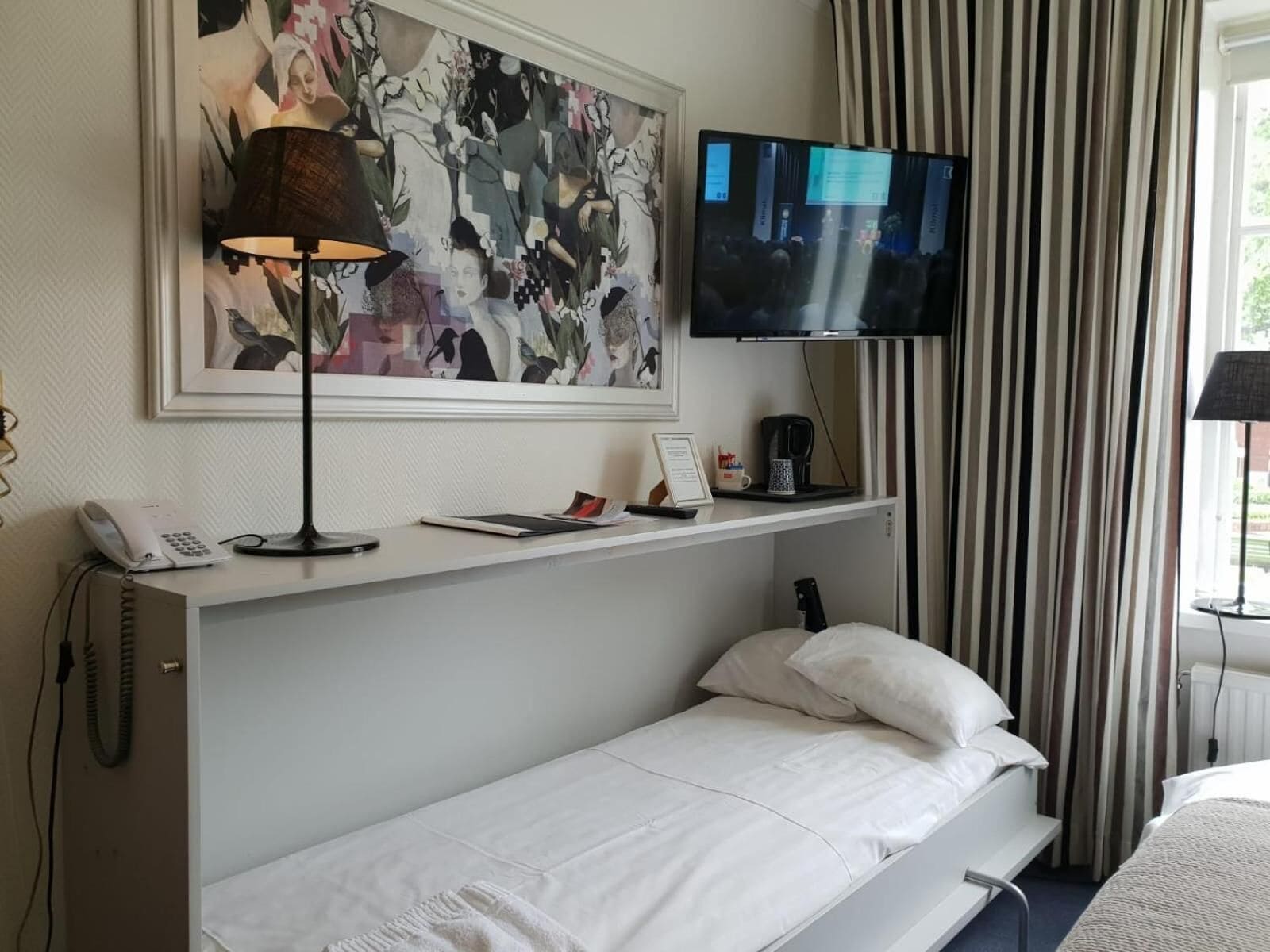 Ett hotellrum med en säng, ett skrivbord med en lampa, en tv och en tavla på väggen.