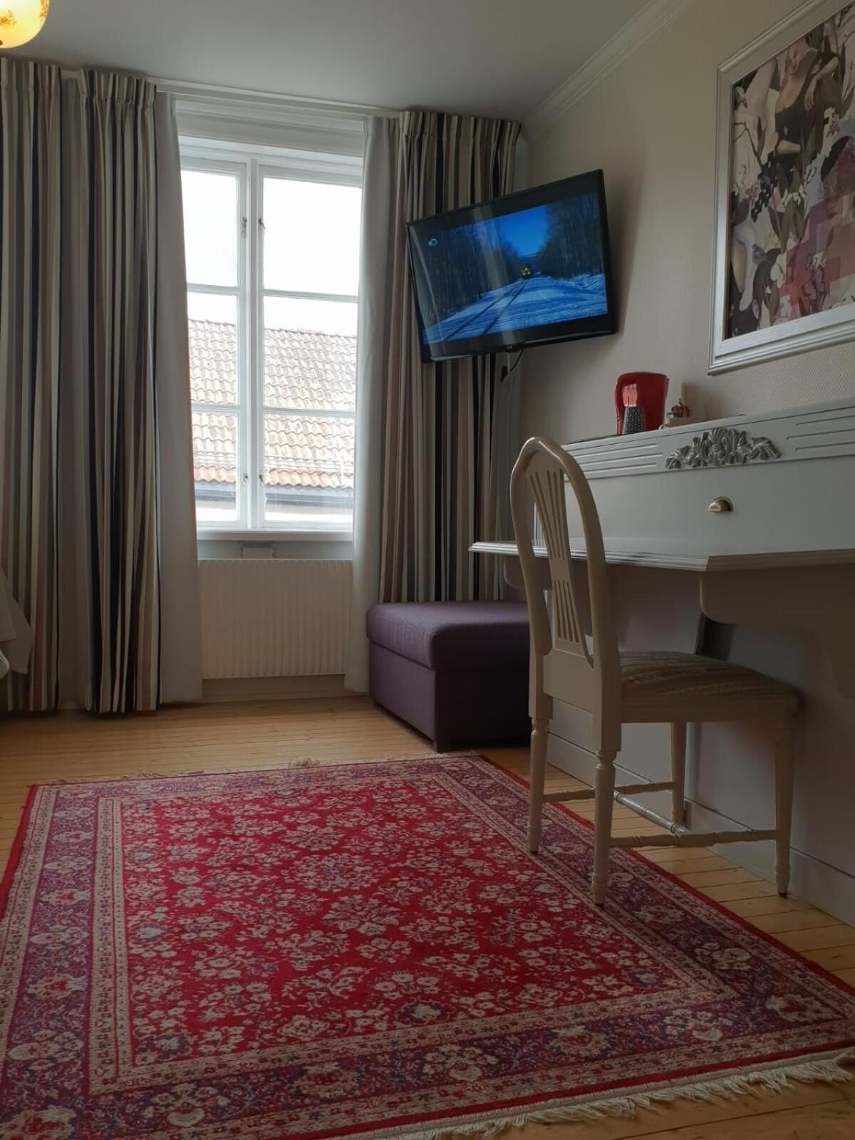 Una habitación con una televisión montada en la pared, un escritorio con una silla, un reposapiés morado, una alfombra roja estampada y una ventana con cortinas.