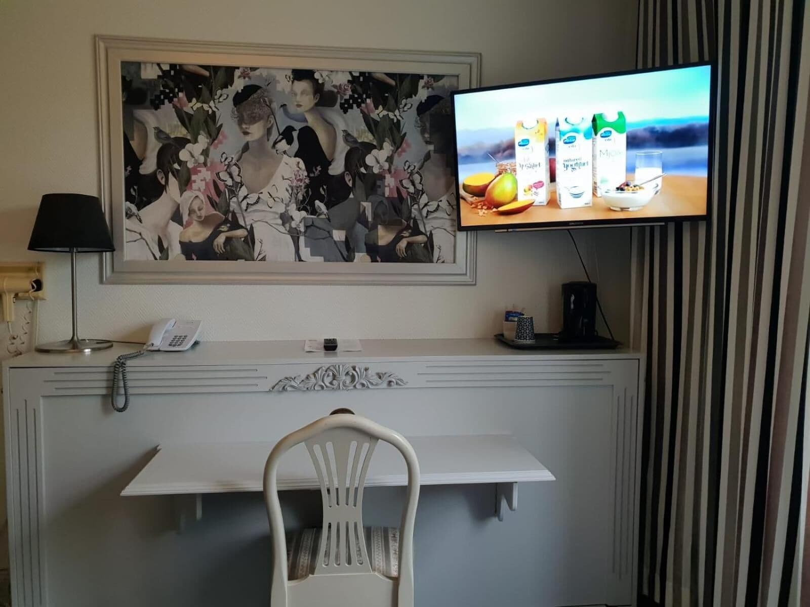 Habitación de hotel con un escritorio, una silla, un teléfono, una cafetera, un televisor mostrando cartones de jugo y un cuadro grande con motivos florales en la pared.