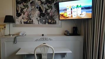 Habitación de hotel con un escritorio, una silla, un teléfono, una cafetera, un televisor mostrando cartones de jugo y un cuadro grande con motivos florales en la pared.