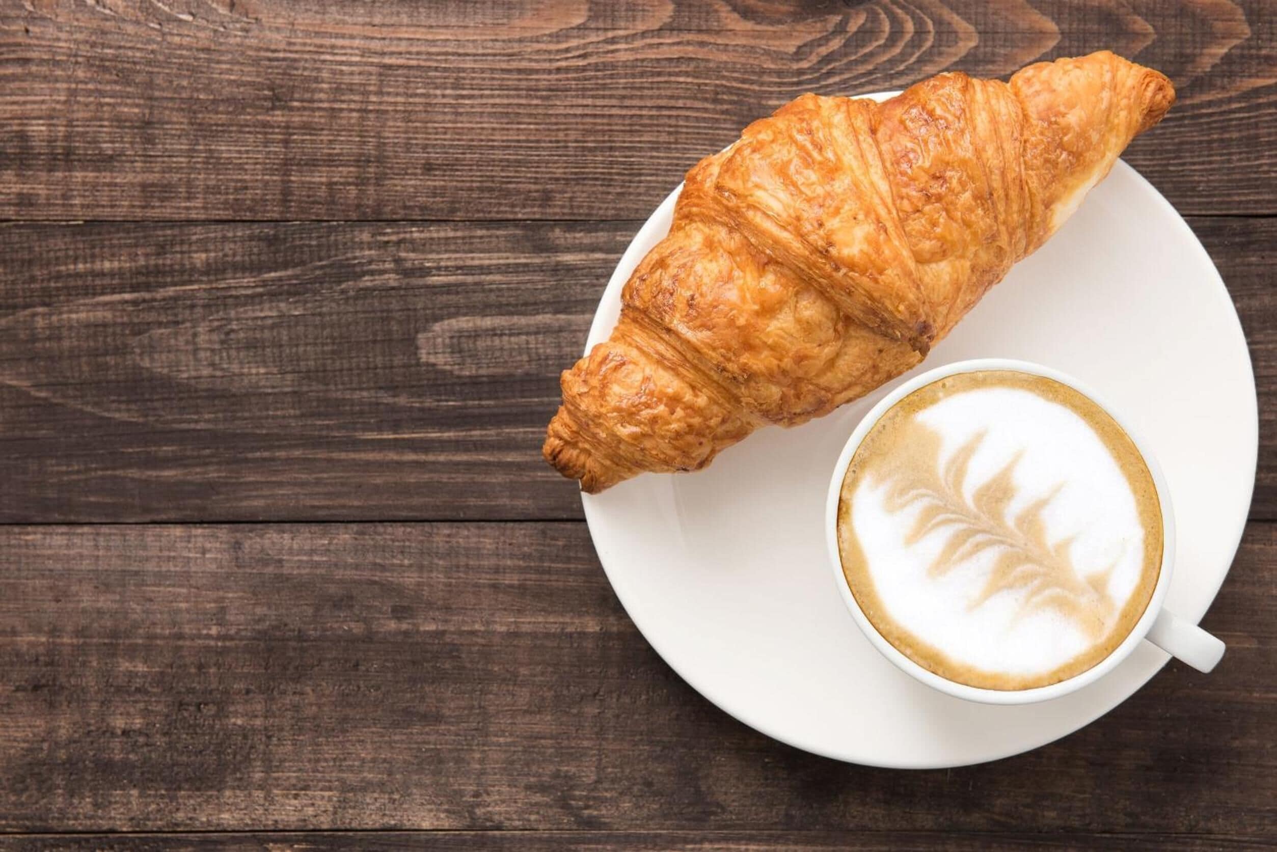 A croissant and a cup of coffee on a white plate, placed on a wooden table.