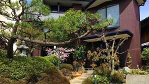 A traditional Japanese house with a stone staircase leading to a wooden deck, surrounded by well-maintained gardens and a large tree. - Hananobou Kinkakuji-michi (Kyoto)