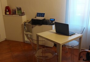Un piccolo ufficio con scrivania, computer portatile, stampante e sedia trasparente in plastica.