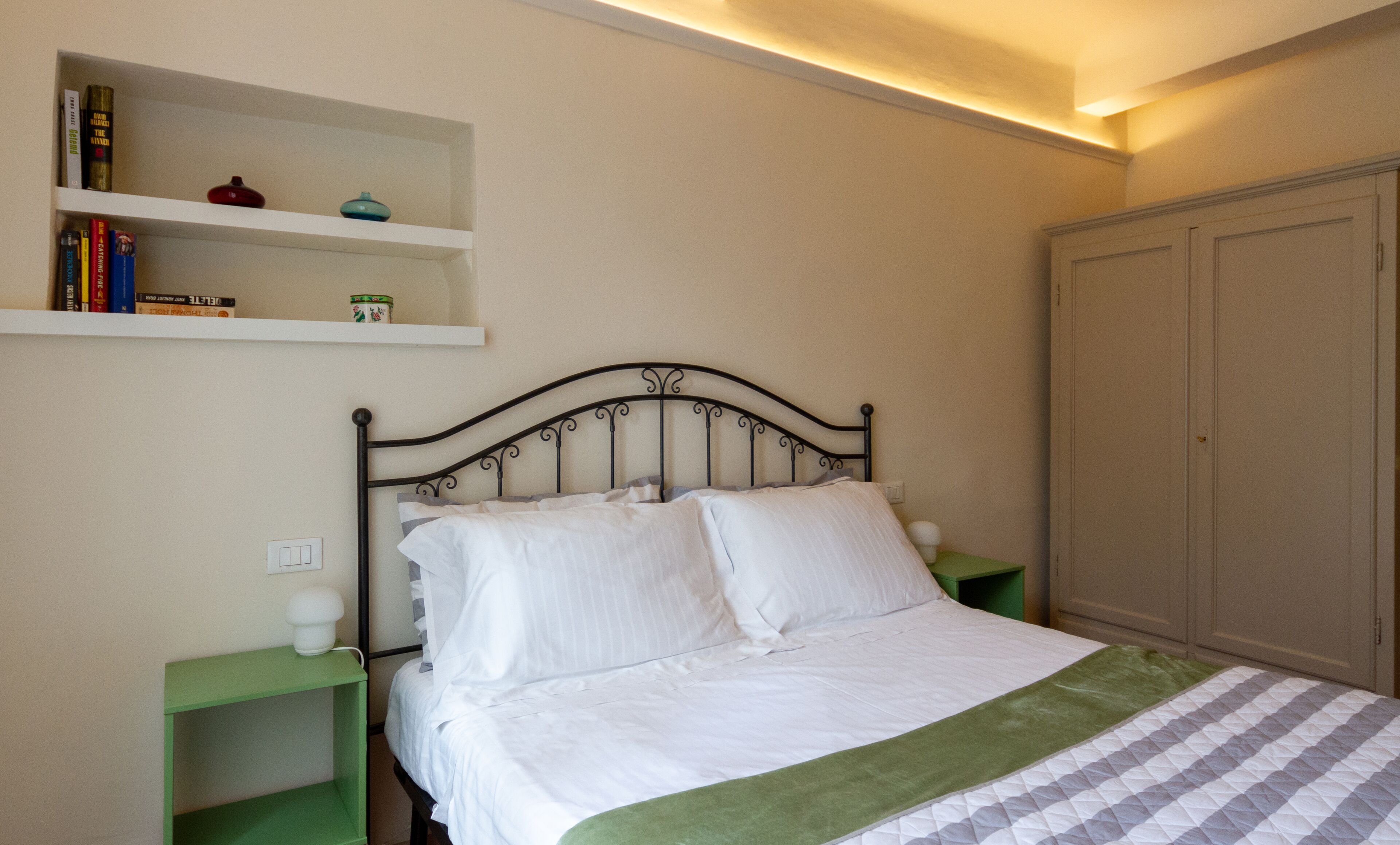 A neatly made bed with white linens and a green blanket, a metal headboard, a bedside table with a lamp, and a shelf with books and decorative items.