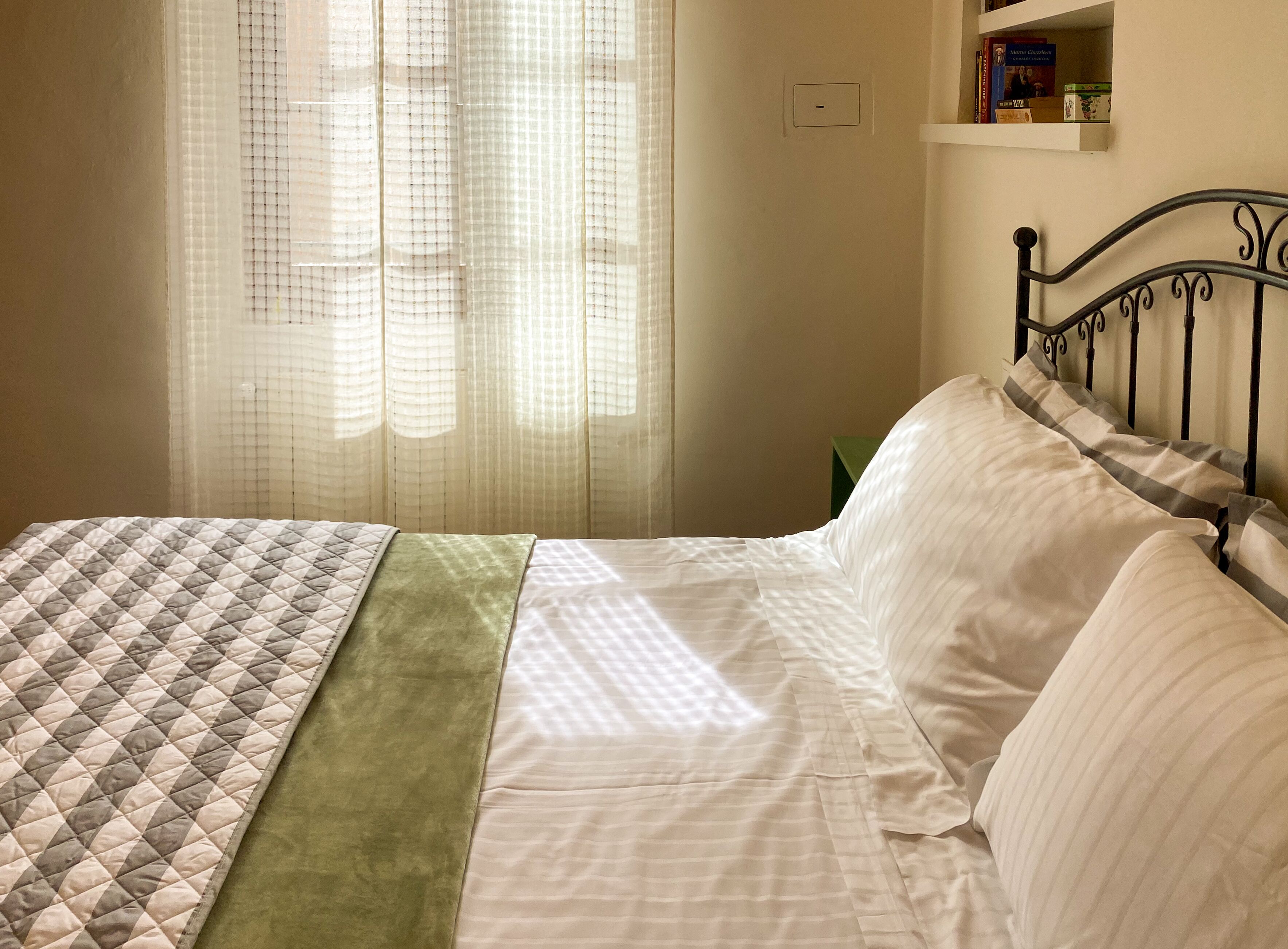 A neatly made bed with a quilt and a green throw blanket, a metal headboard, and a shelf with books and a box.