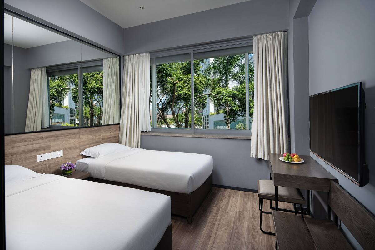 A hotel room with two beds, a desk, a TV, and large windows with curtains.