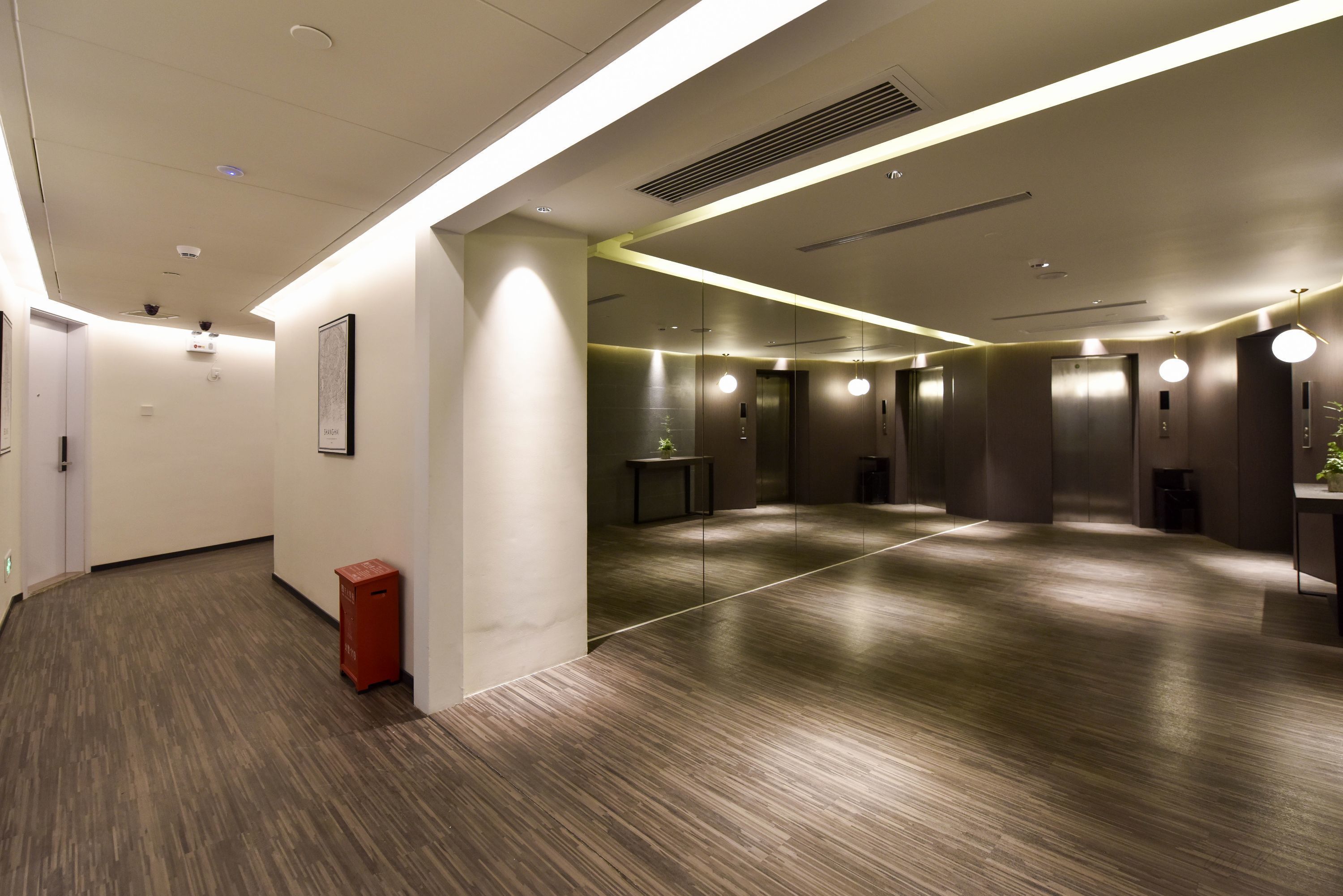 A modern hotel lobby with a wooden floor, a red trash bin, and a large mirror reflecting the space.