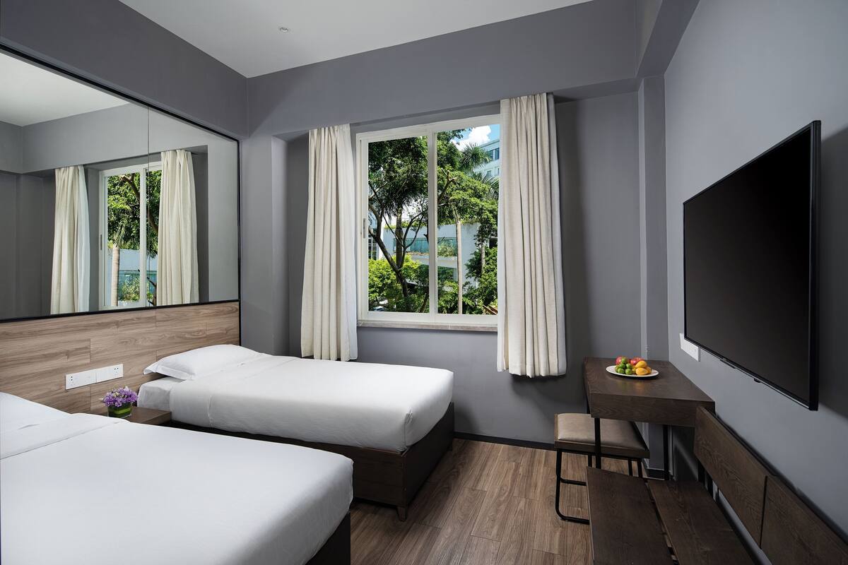 A modern hotel room with two beds, a desk, a chair, a TV, and a window with curtains.