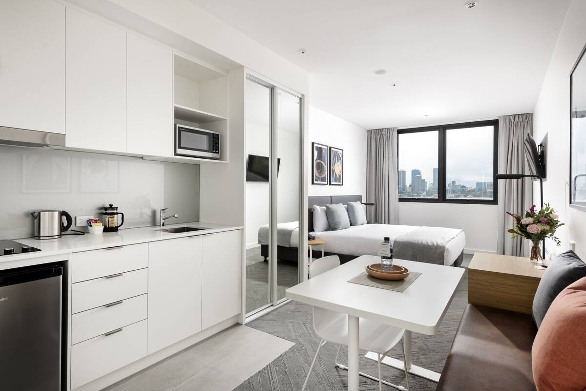 A modern hotel room with a kitchenette, a bed with gray bedding, a small table with a vase of flowers, and a city view through large windows.