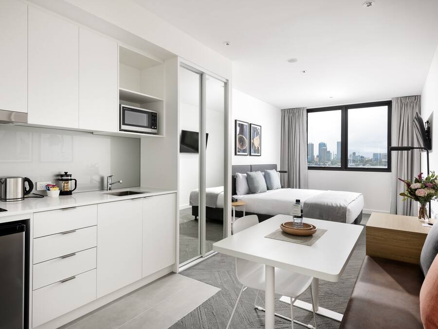 Una habitación de hotel moderna con kitchenette, una cama con ropa de cama gris, una mesita con un florero y vistas a la ciudad a través de amplios ventanales.