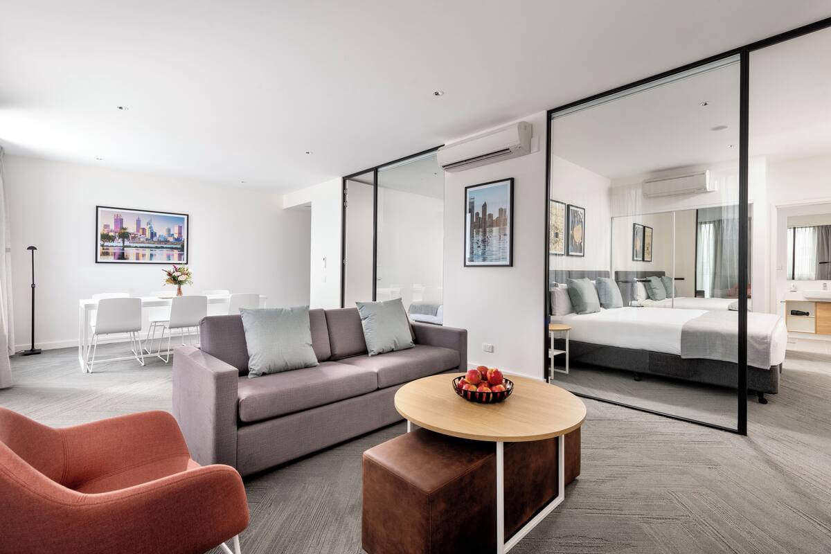 A modern hotel room with a grey sofa, a round wooden coffee table, a red chair, a bed with grey bedding, and a large mirror.