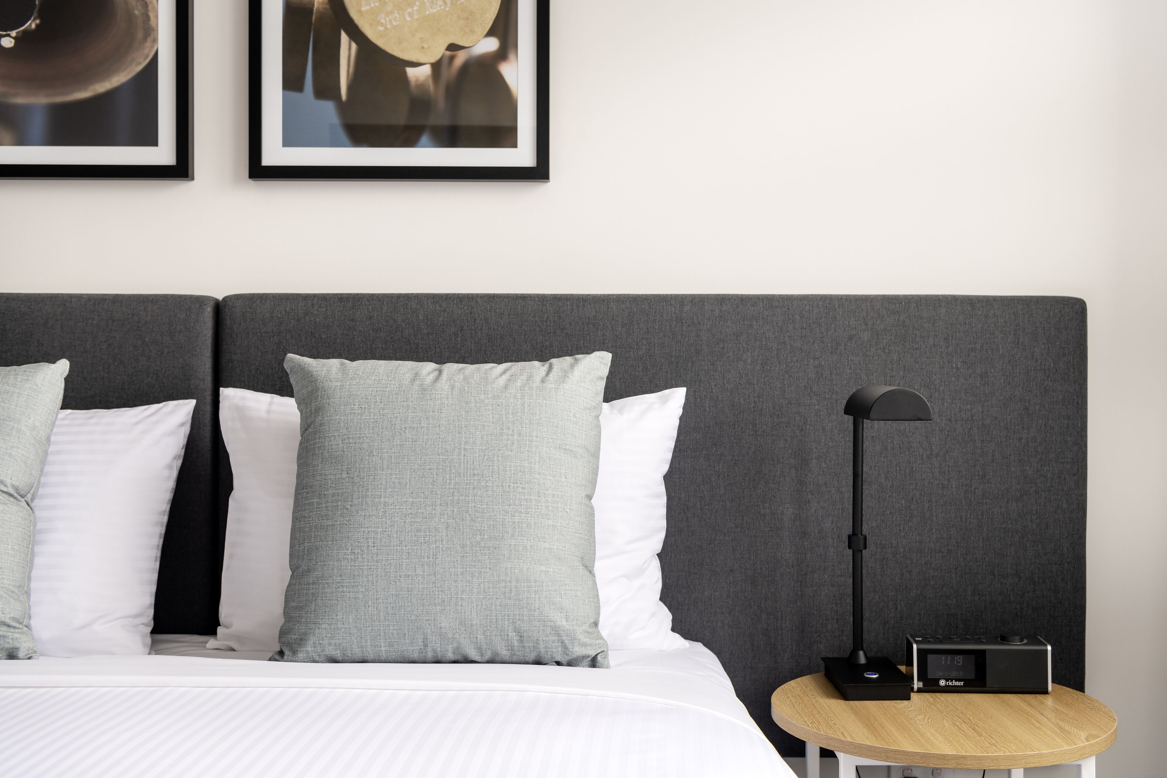 A neatly made bed with white and grey bedding, a round wooden side table with a lamp and radio, and two framed pictures on the wall.