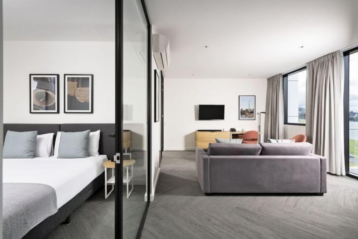 A modern hotel room with a bed, a sofa, a desk, and a large window offering a city view.